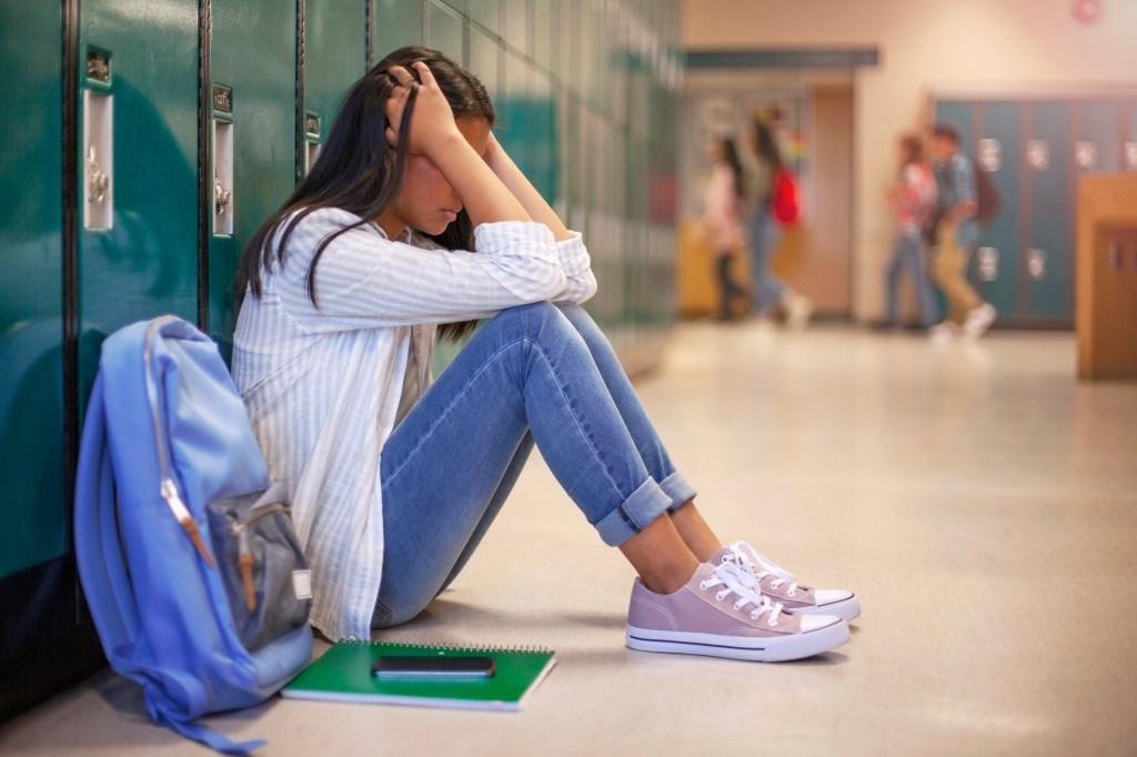 Frustrated Asian teenage girl sitting with head in hands in well-lit school corridor against metal lockers